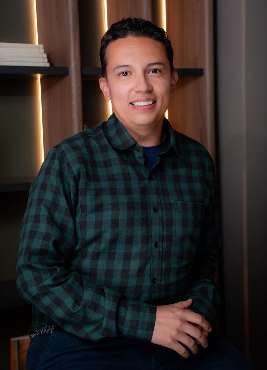 Christian F. Rojas, seated in front of a lit bookshelf, wearing a green checked shirt and smiling at the camera.